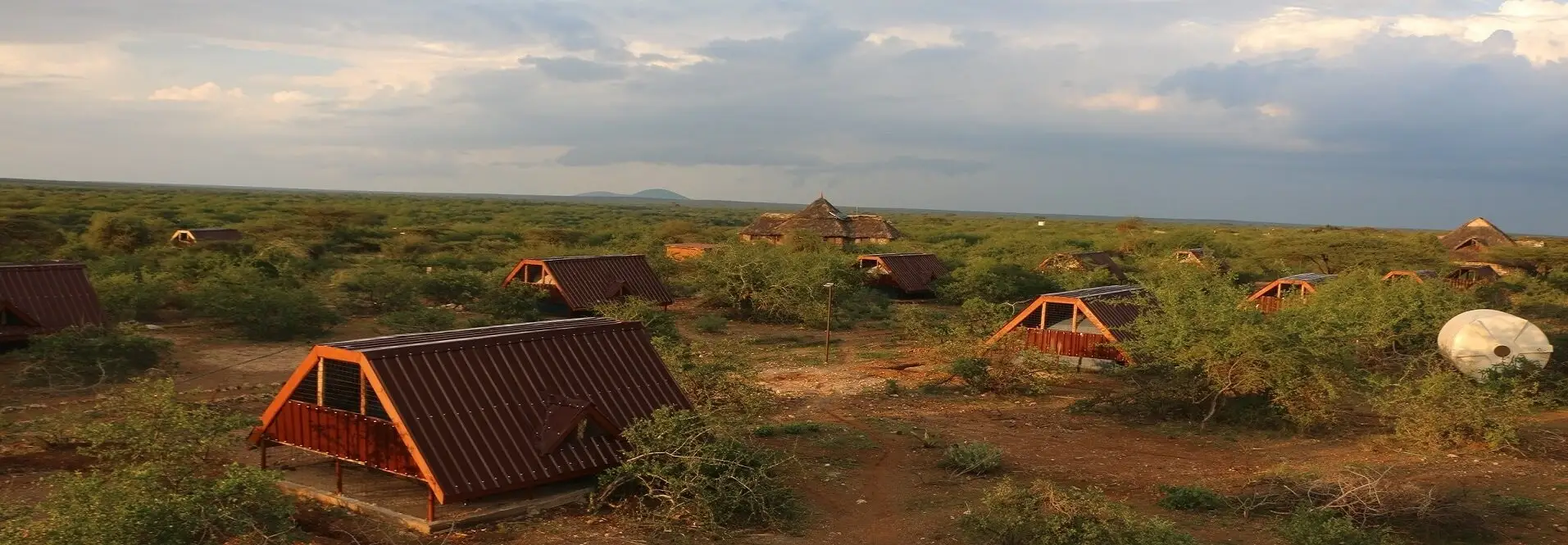 A wide view of the camp under a stormy evening sky, with red-roofed A-frame tents scattered across flat green bush and a thatched building in the distance.