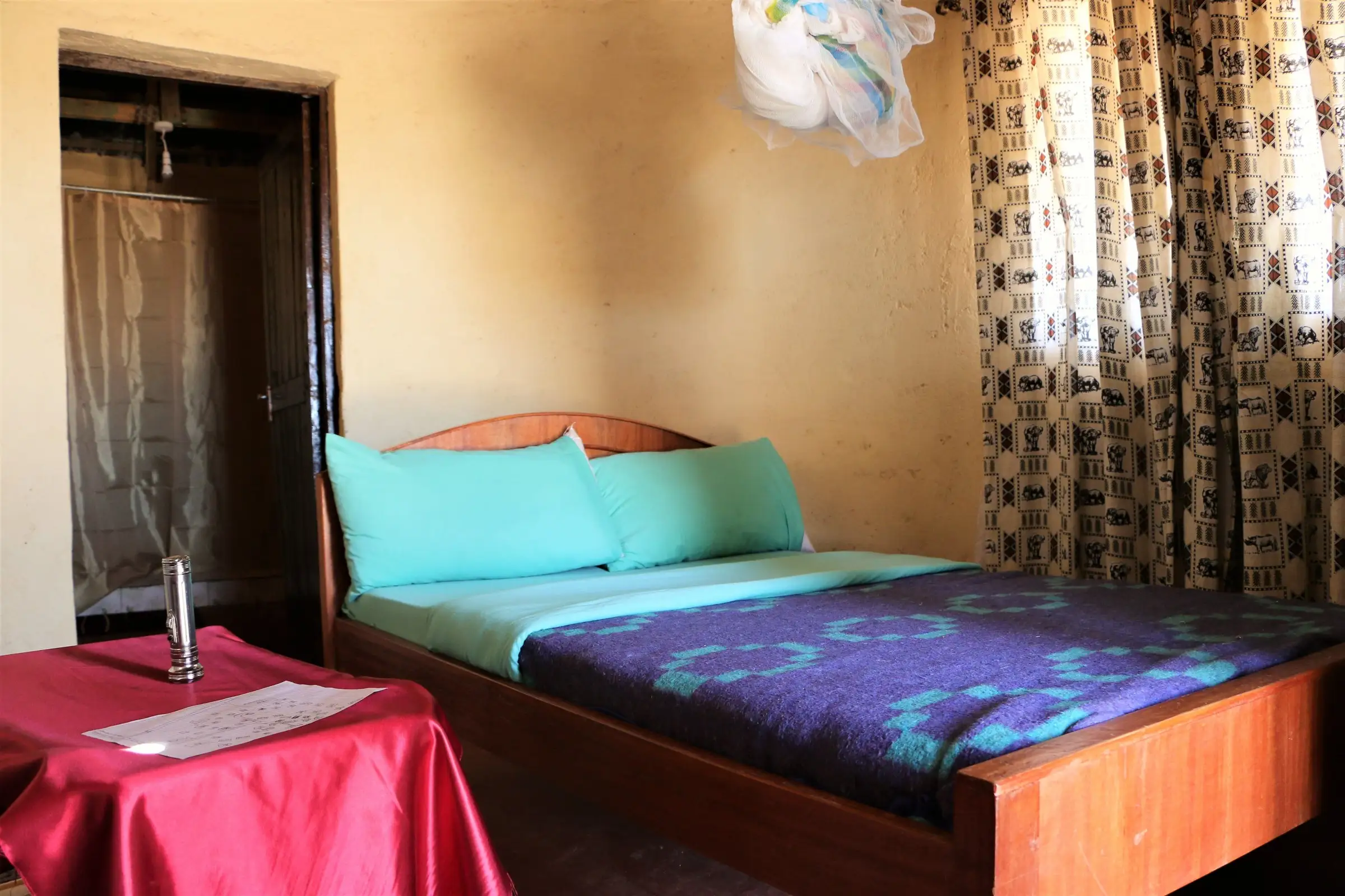 A bedroom with a double bed, teal pillows, a purple and teal blanket, a mosquito net above, animal-print curtains, and a shower curtain behind an open door.