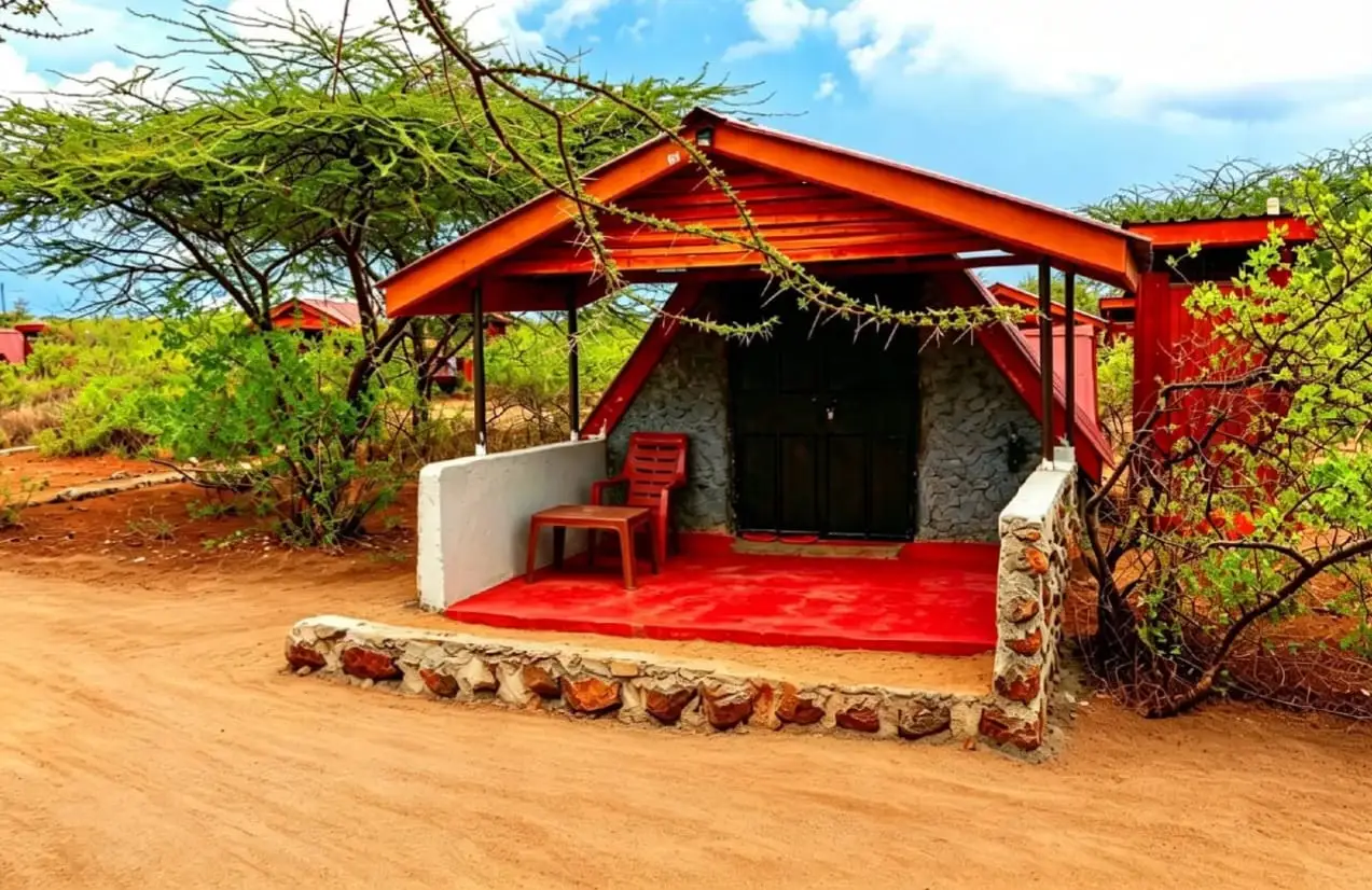 A small cottage with a red roof, grey stone front, white porch wall and a plastic chair and table on a red veranda, among acacia trees and red earth.
