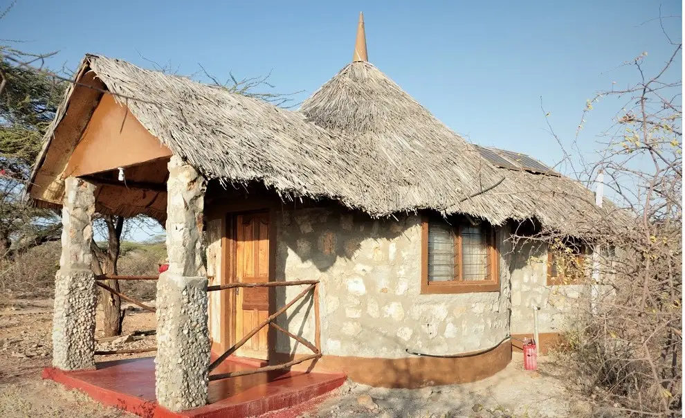 A round stone cottage with a thatched roof, a covered veranda on pebble-faced pillars, a wooden door and a red step.