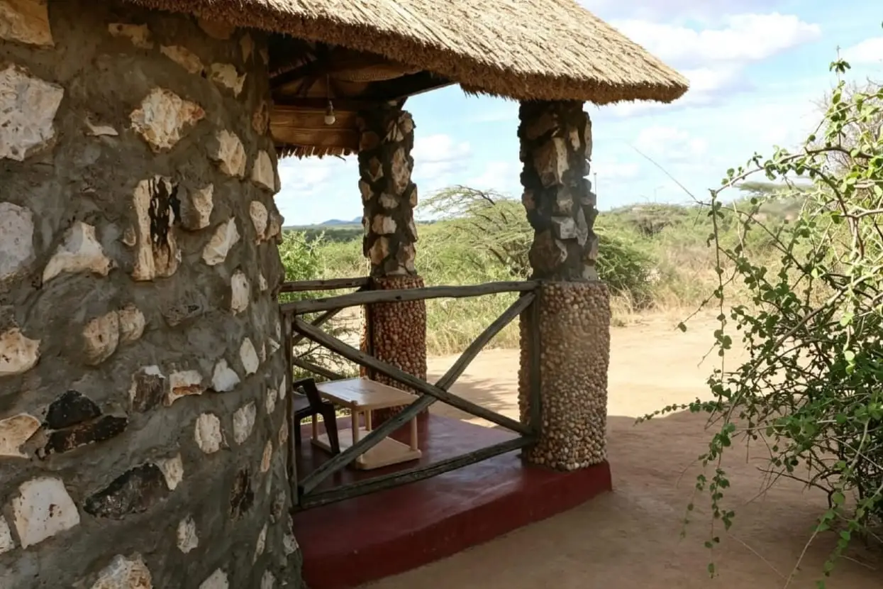 A veranda with a thatched roof, stone and pebble pillars and a rough timber rail, looking out over dry bush and distant hills.