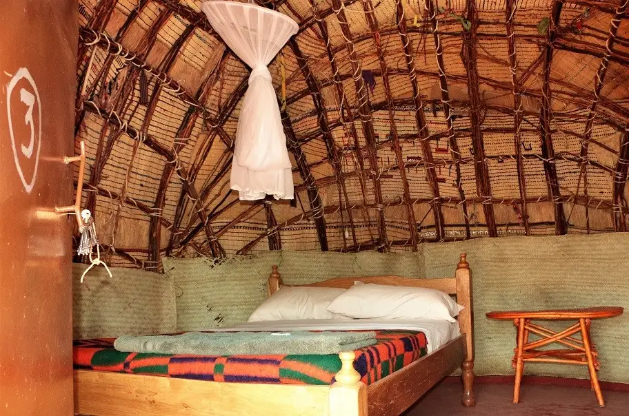 Inside a traditional hut with a domed roof of woven mats and sticks, a wooden bed with a white mosquito net and a striped blanket, and a small wooden stool.