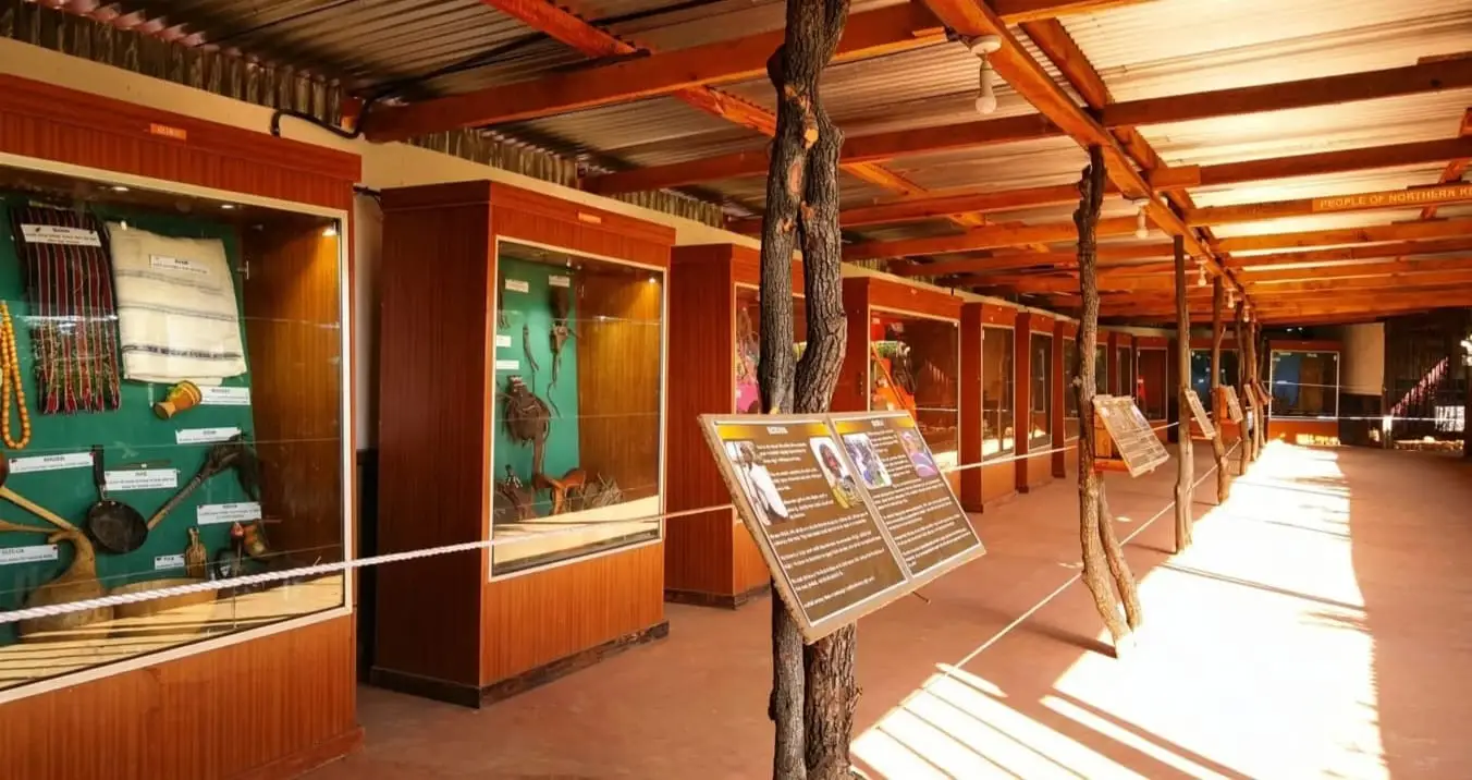 A long hall of wooden display cases holding traditional cloth, beadwork, gourds and tools, with information boards beside a rope barrier.