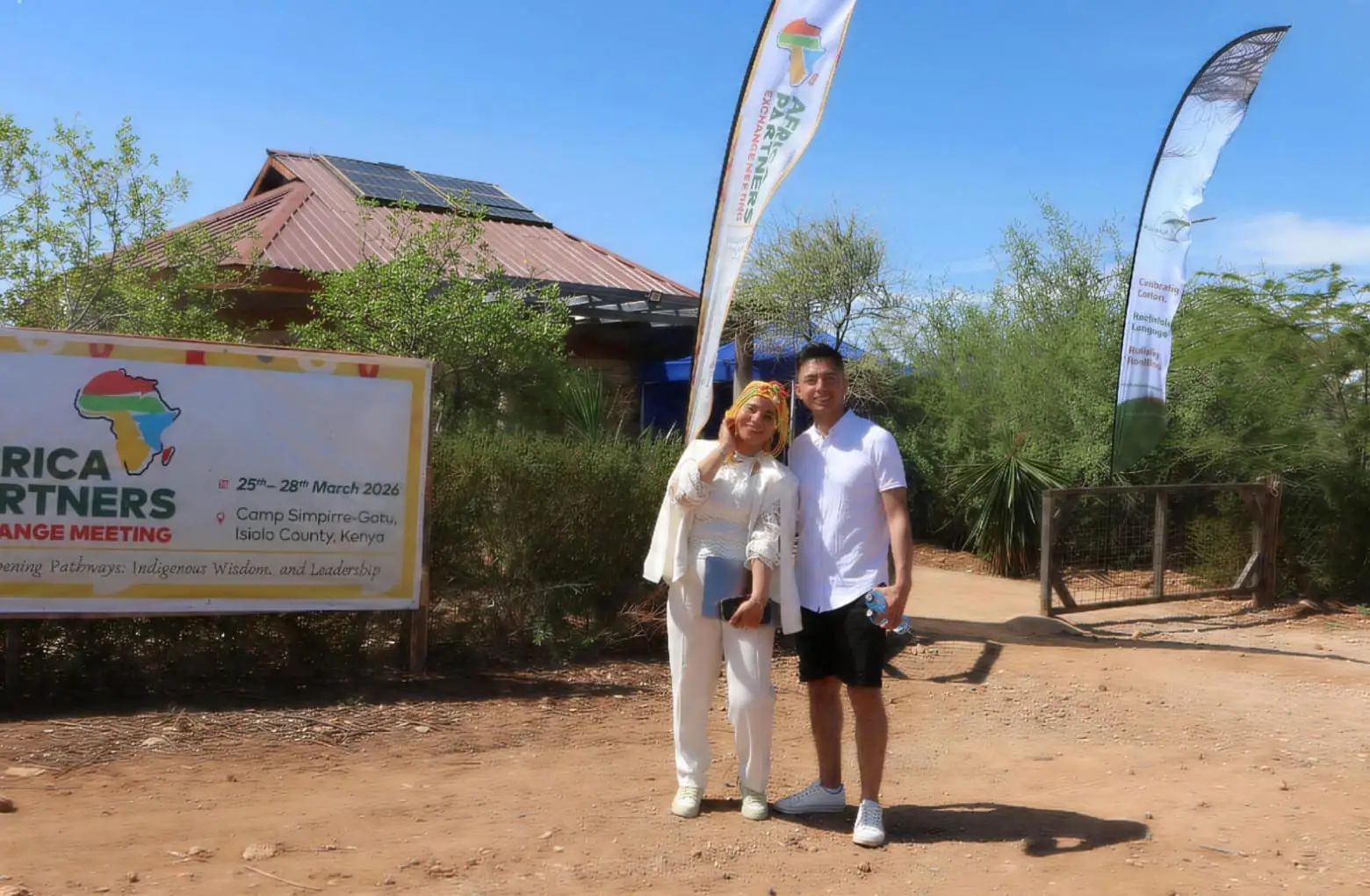 Two people standing in front of an event banner, with a solar-panel roof and tall event flags behind them.