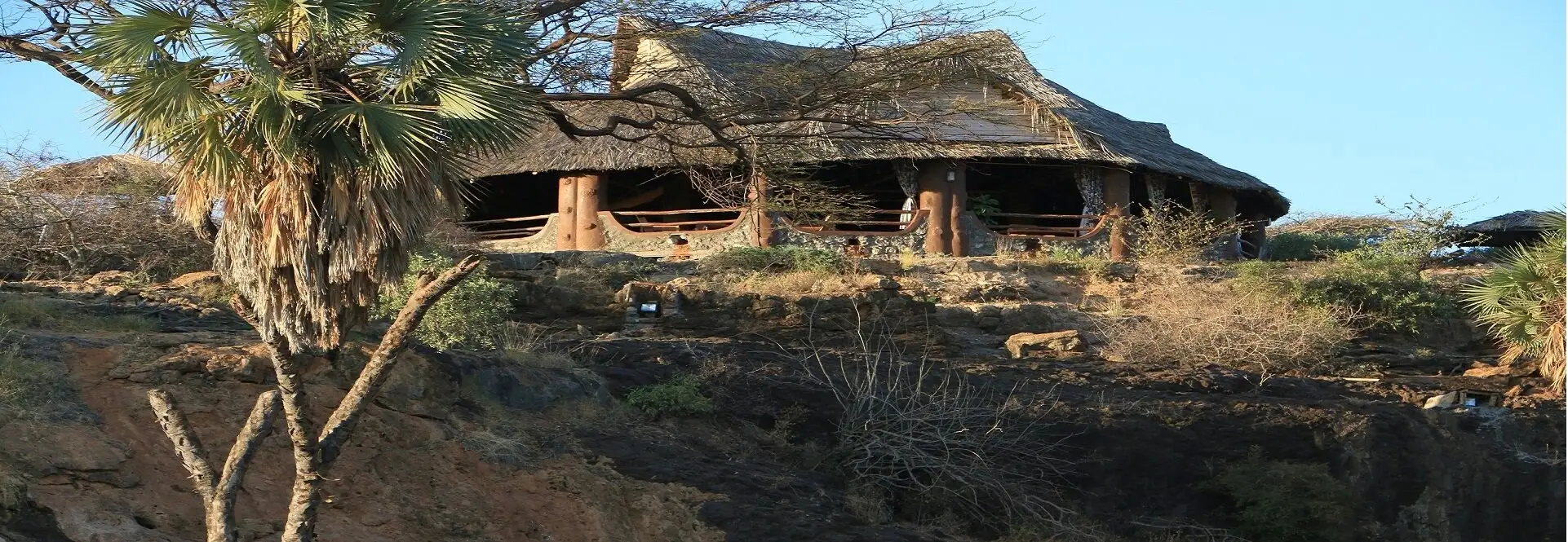 A large thatched lounge with log posts and a stone-and-timber rail, sitting on a rocky ridge above dark rock and a palm tree, under a clear blue sky.