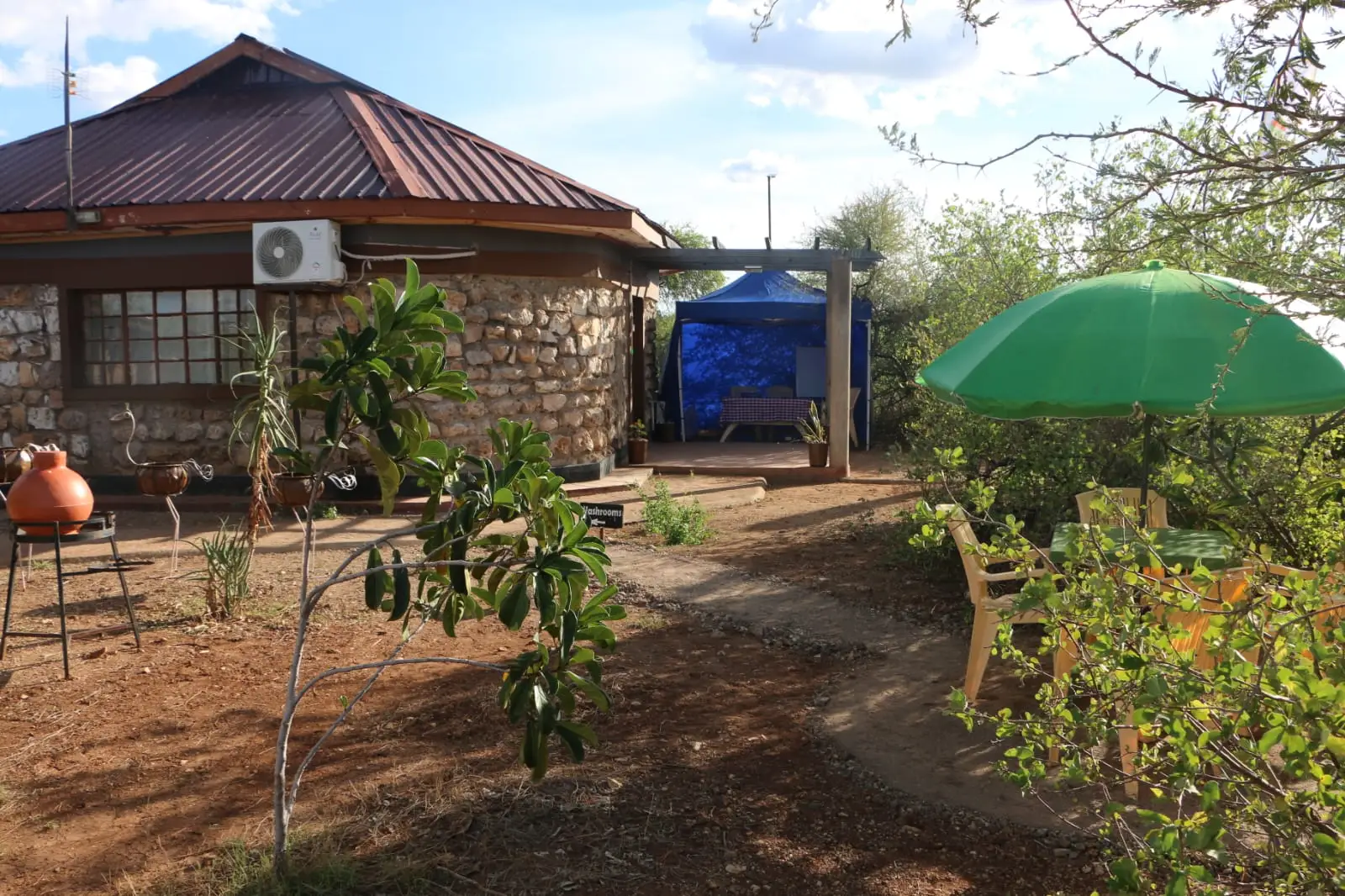 A round stone house with a tin roof and air conditioner, a garden path, a clay pot, a green umbrella over a table and chairs, and a blue gazebo beyond.