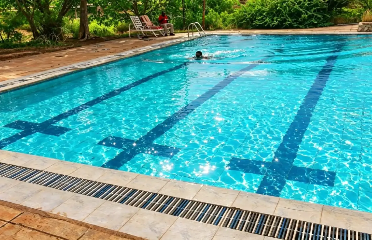 A clear blue swimming pool with dark lane markings. A swimmer is in the water and a guest sits on a lounger under acacia trees.