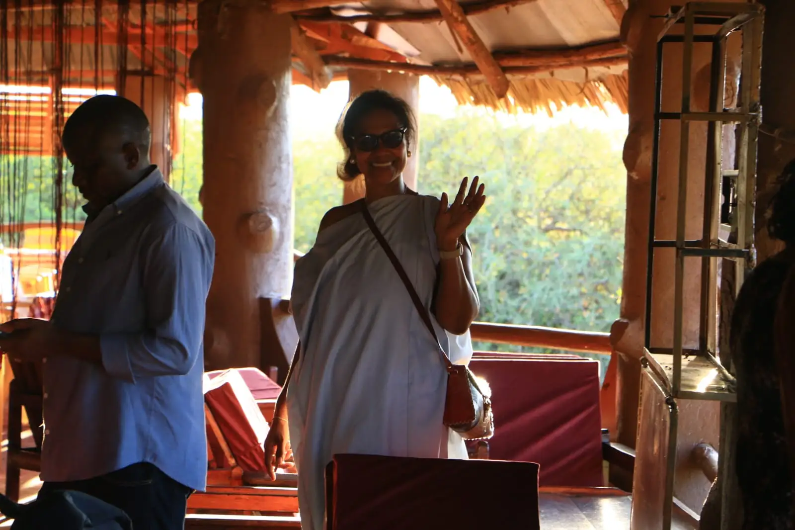 Inside a thatched, open-sided lounge with log posts and red cushioned seats, a smiling guest waves while another person looks at a phone.