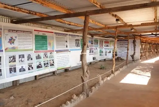 A covered museum corridor with printed boards about East African communities along one wall, a rope barrier and a sunlit path.