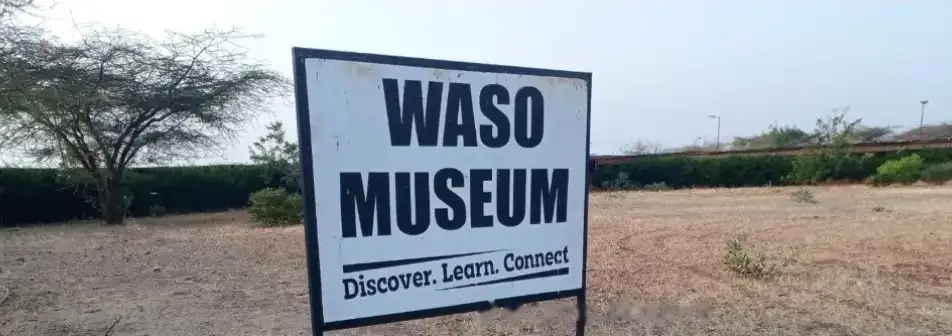 The white Waso Museum sign, with the line 'Discover. Learn. Connect.', standing in dry grass beside a hedge.