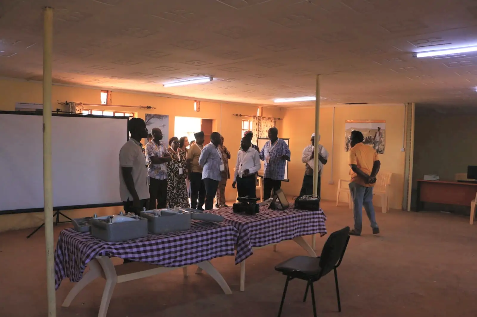 A workshop room with a projector screen, two tables with checked cloths holding equipment cases, and a group of people standing and talking.